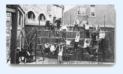 Large group of boys playing on a junglegym at rear of old school building.