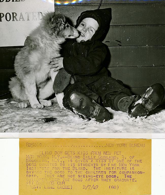 1947 image of winter dressed child sitting in the snow being kissed by his dog. 